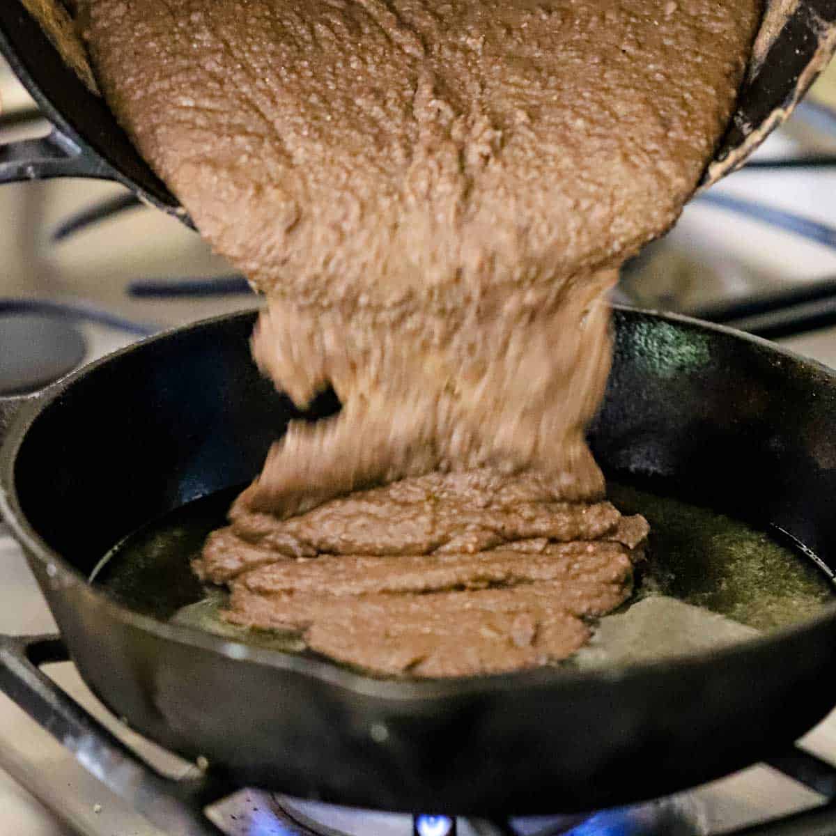 A person transferring puréed pinto beans into a cast-iron skillet that contains melted pork lard on a gas stove.