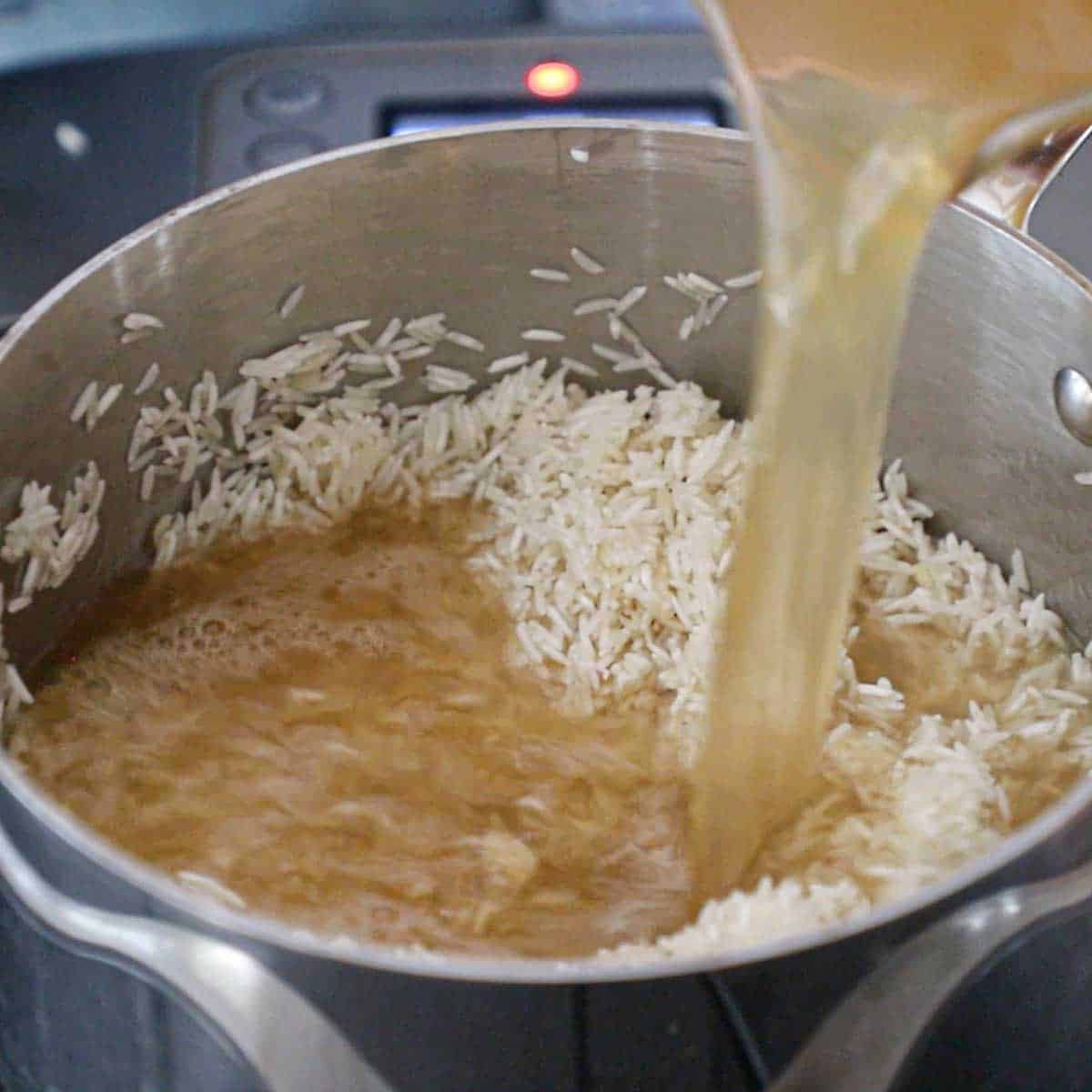 A person pouring chicken broth from a glass measuring cup into a saucepan filled with toasted Jasmine rice.