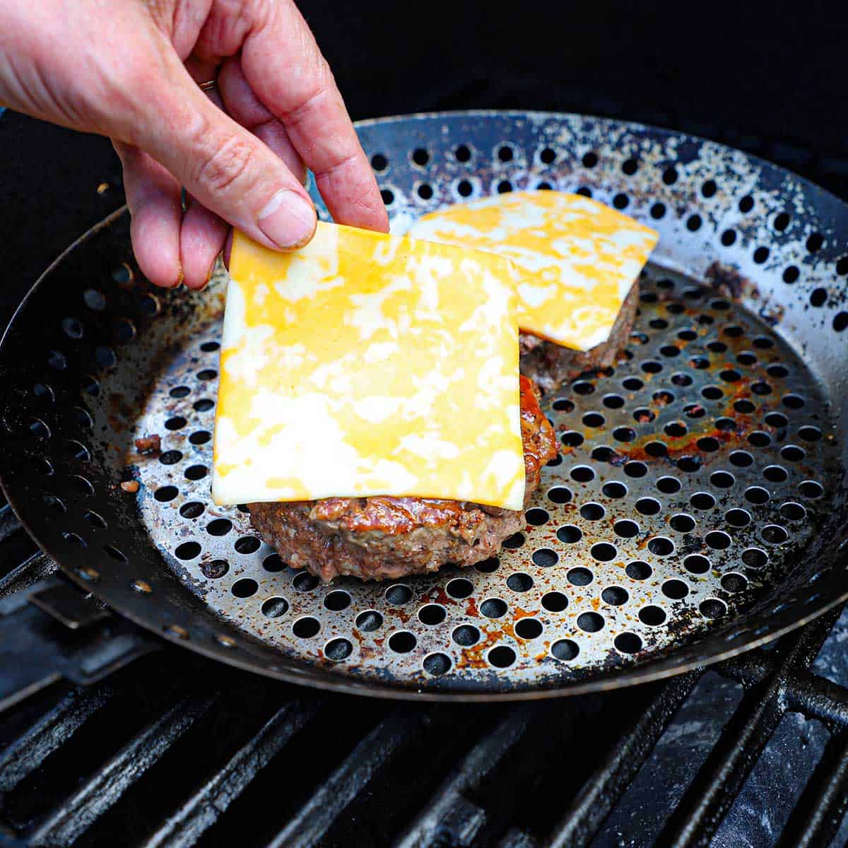A person placing slices of Colby Jack cheese over two burger patties that have been cooked on a grill pan on a gas grill.
