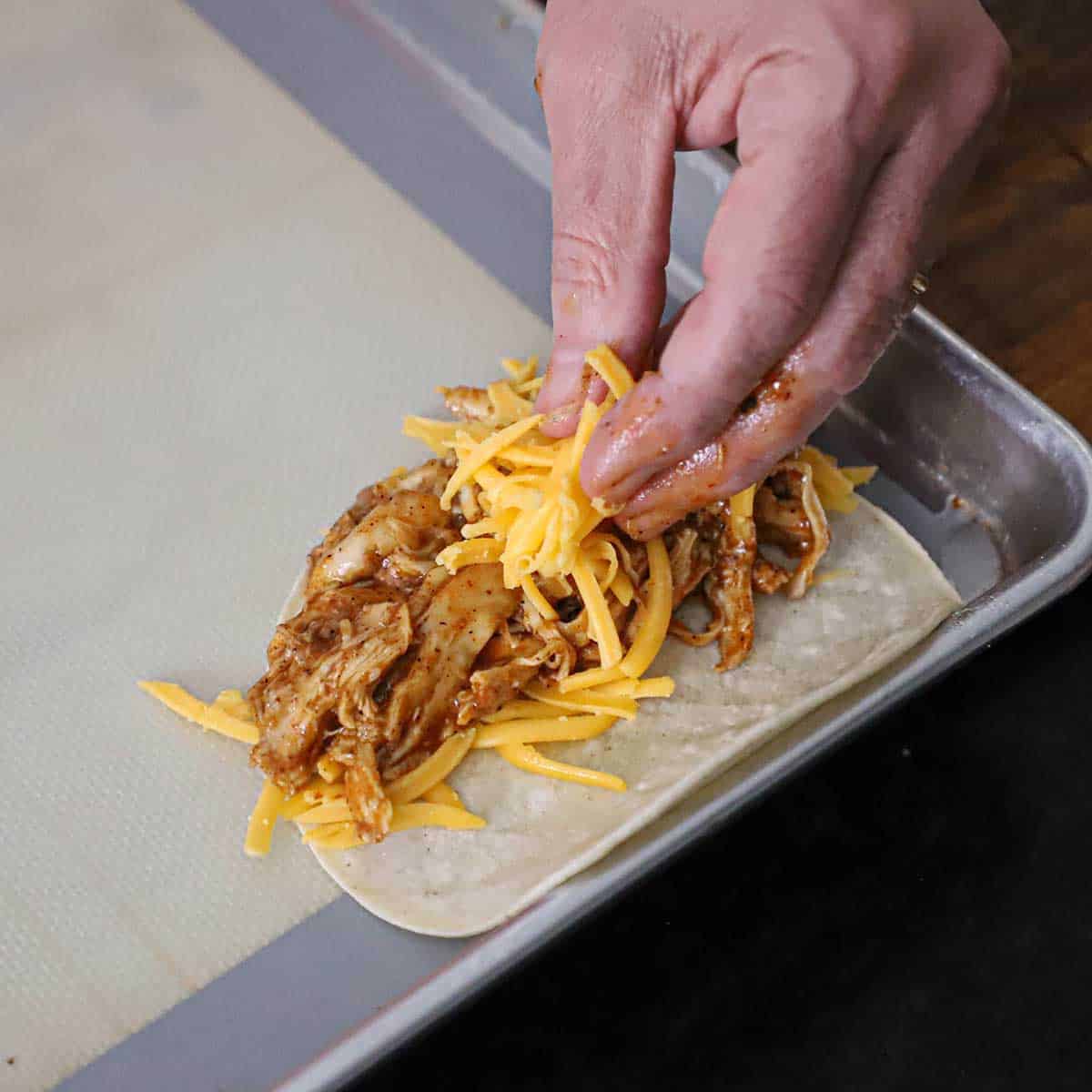 A person placing shredded cheddar cheese over red enchilada shredded chicken resting on more cheese all on one half of a corn tortilla.