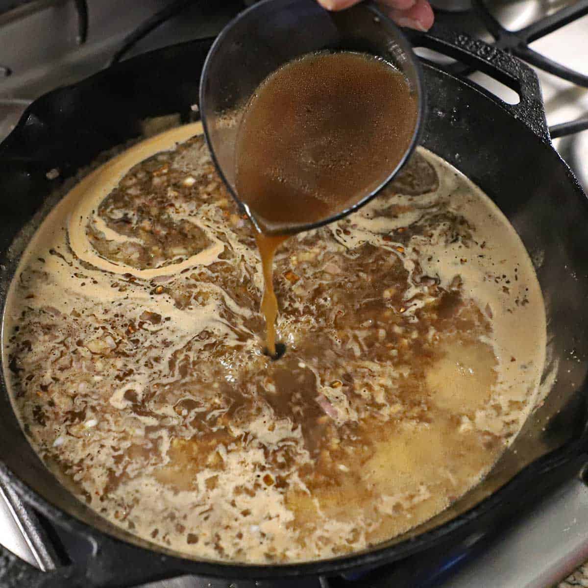 A person pouring beef stock from a measuring cup into a large cast-iron skillet that is filled with a simmer beef stock and Madeira wine sauce.