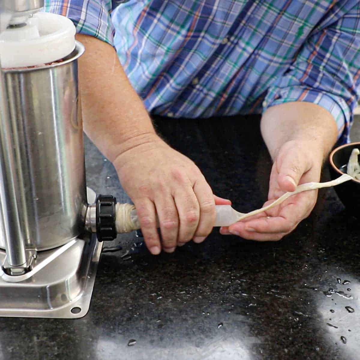 A person scrunching a long hog casing onto a tube that is attached to a metal canister used for stuffing sausage.