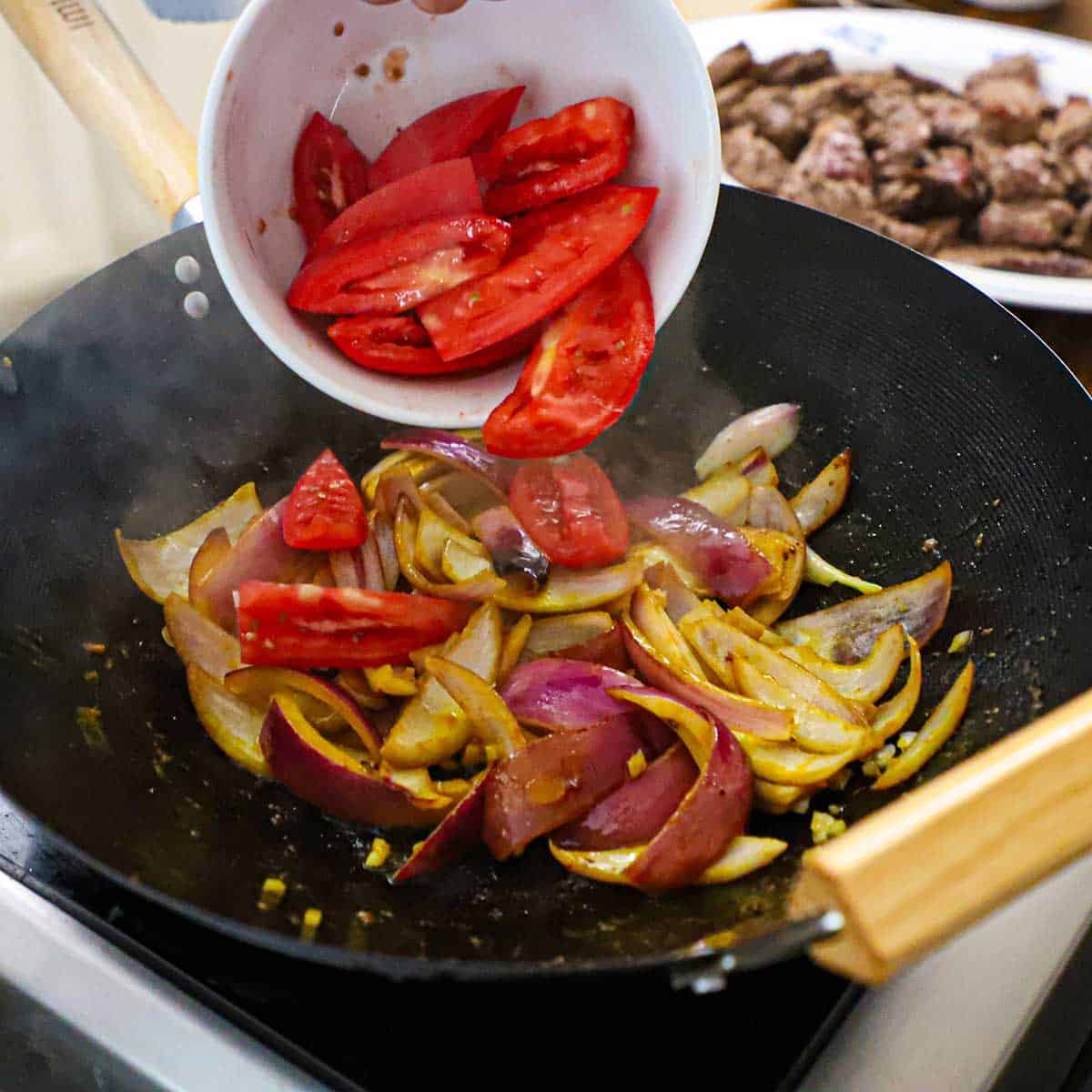 A person transferring quartered Roma tomatoes into a wok that is filled with red onion wedges, garlic, and pepper paste being stir-fried.
