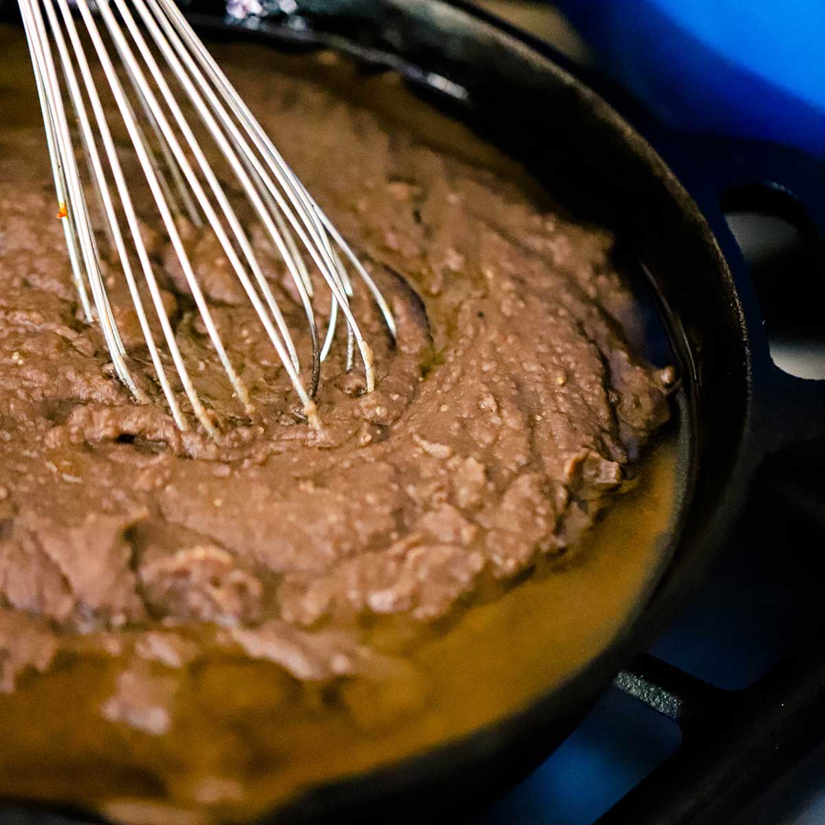 A person using a whisk to combine homemade refried beans in a cast-iron skillet with melted pork lard on a gas stove.