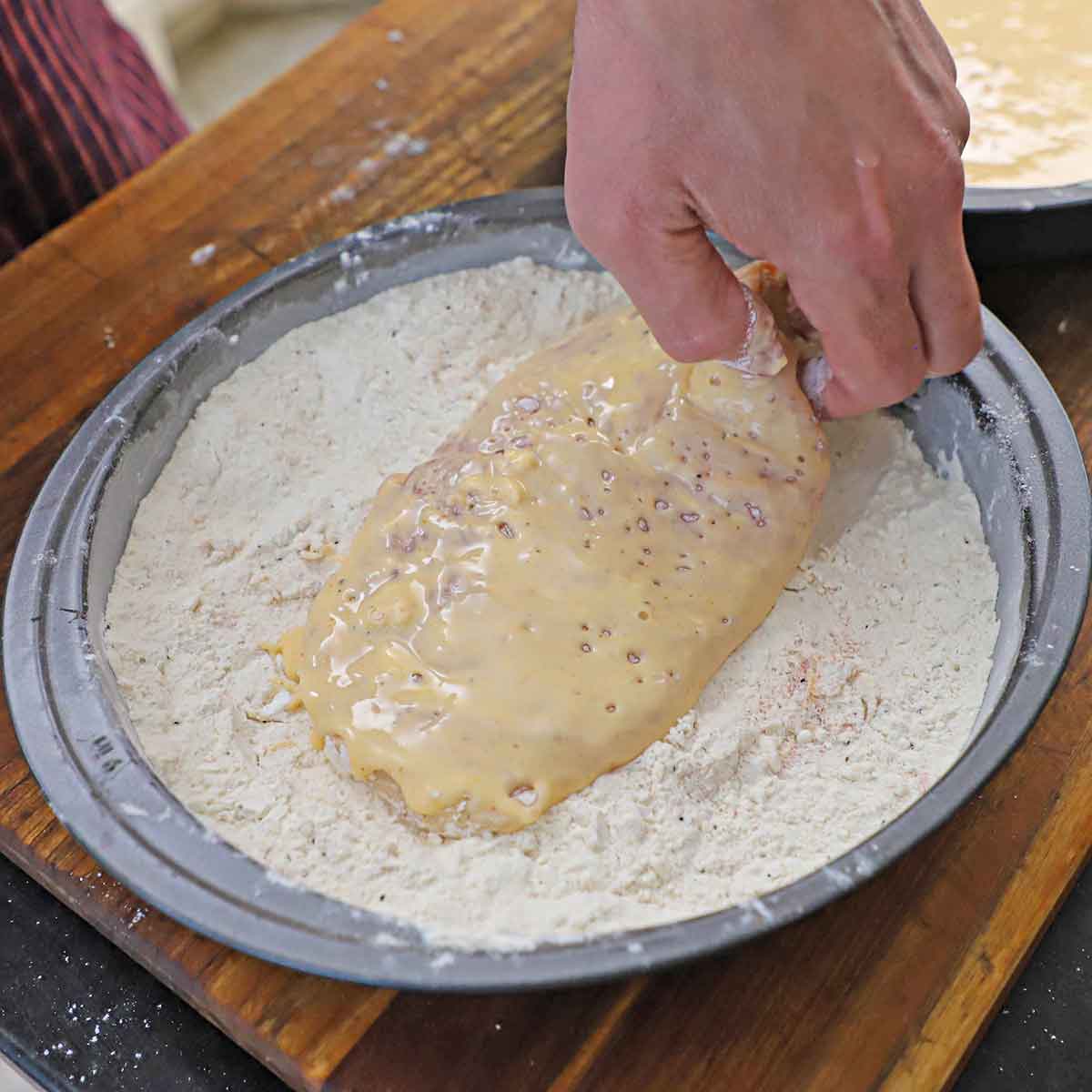 A person dredging a thin chicken breast cutlet that is coated with a wet batter in a pan filled with seasoned flour.