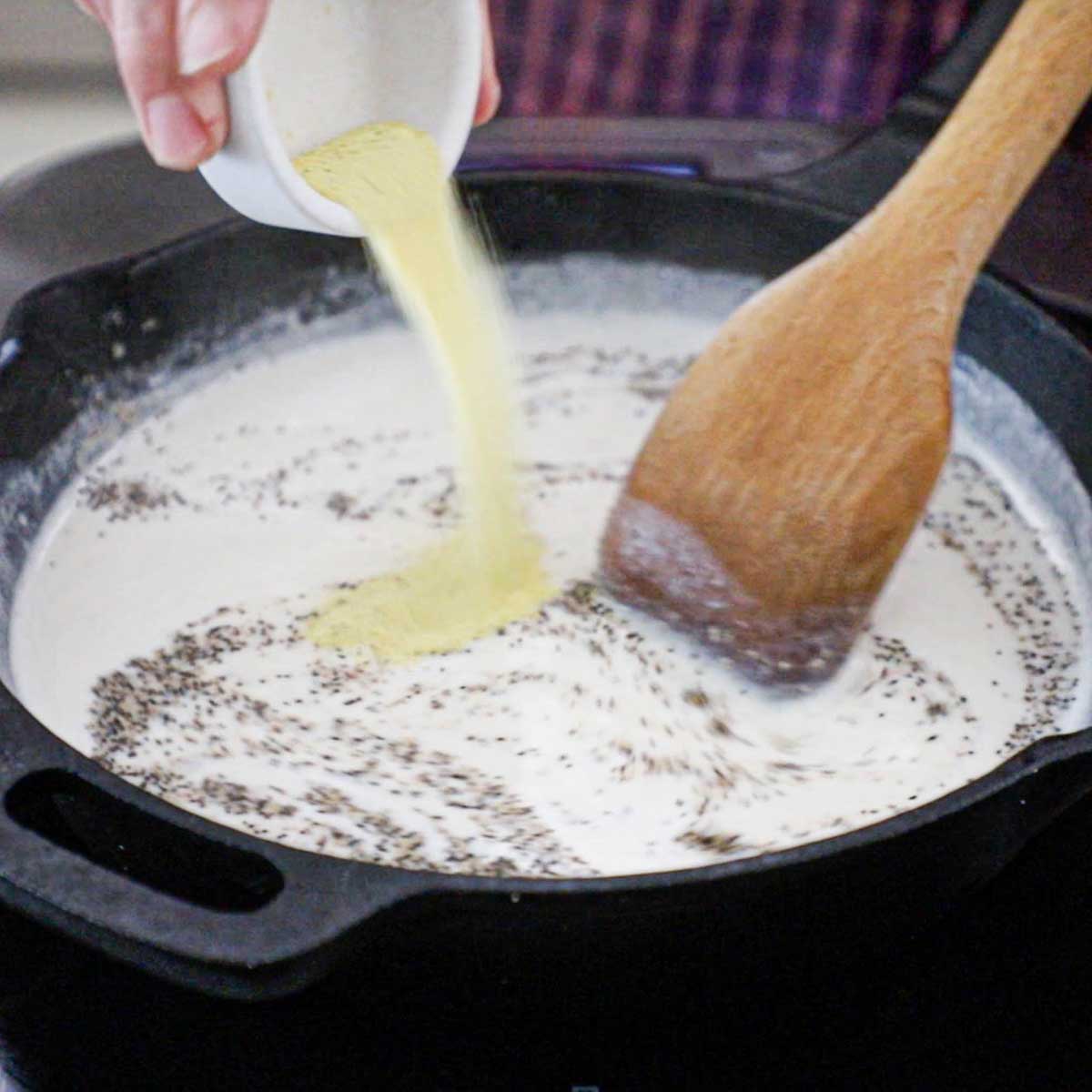 A person adding adobo seasoning into a cast-iron skillet that is filled with freshly prepared classic Southern cream gravy with black pepper.