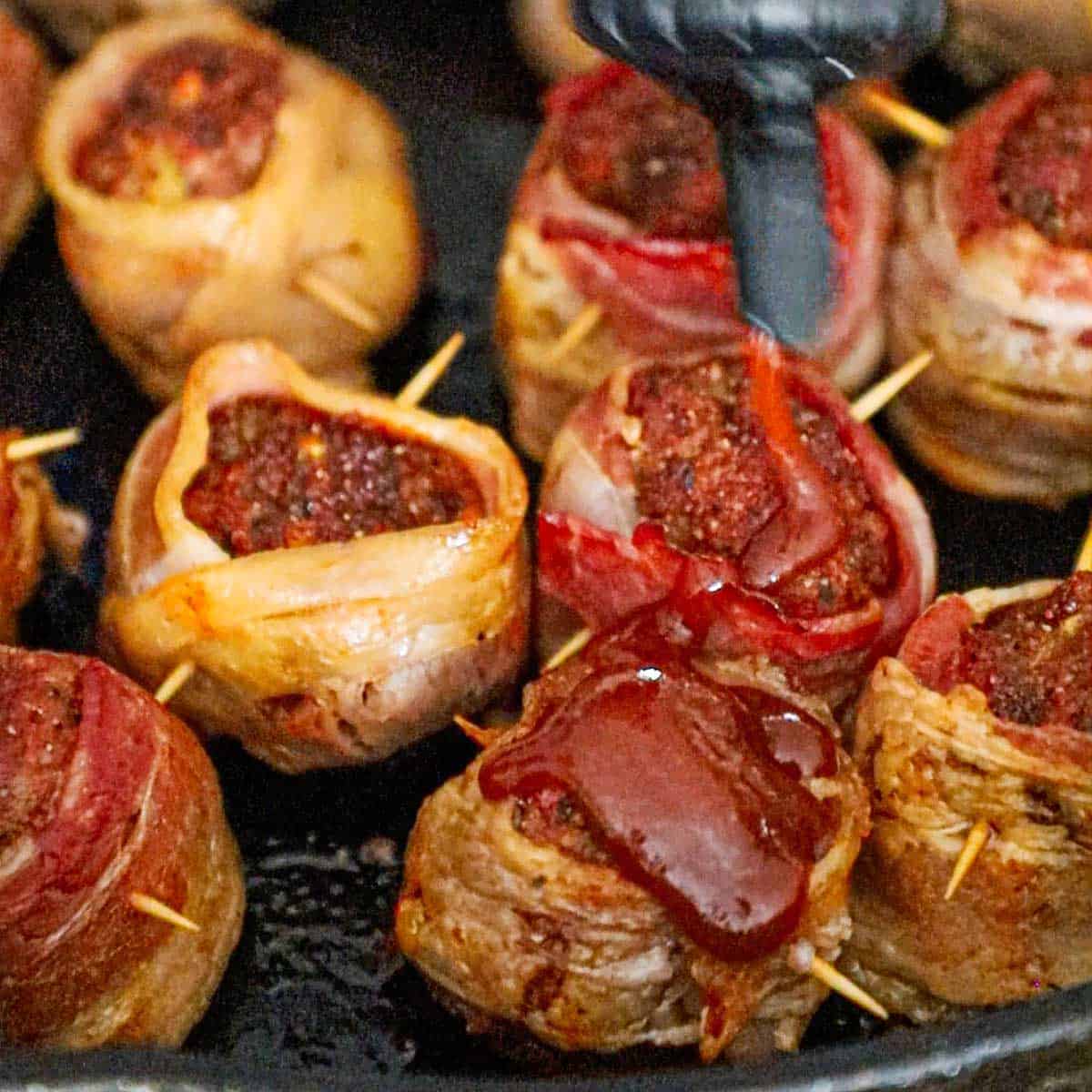 A person drizzling BBQ sauce over the top of smoked bacon-wrapped meatballs that are partially cooked and resting in a large cast-iron skillet.