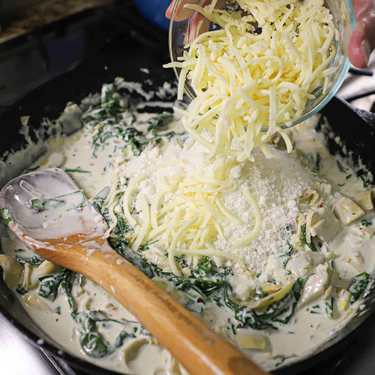 A person transferring shredded Mozzarella cheese from a glass bowl into a cast-iron skillet that is filled with a simmering Spinach and Artichoke Dip sauce.