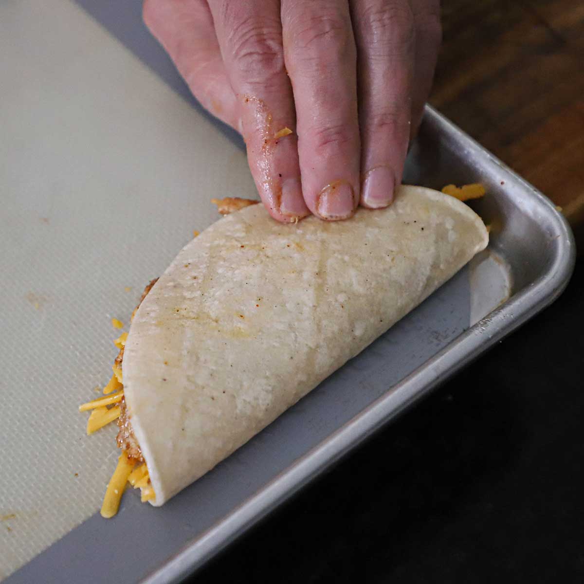 A person folding one half of a white corn tortilla over a filling of red enchilada chicken and shredded cheddar cheese.
