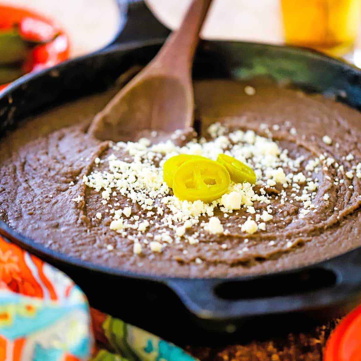 A close-up view of a large black cast-iron skillet filled with authentic homemade refried beans topped with crumbled Cotija cheese and three slices of pickled jalapeños.