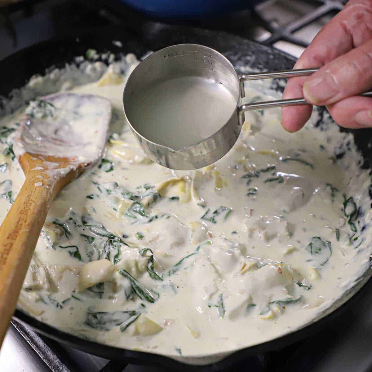 A person pouring a small amount of pasta water from a metal measuring cup into a cast-iron skillet filled with simmering Spinach and Artichoke Dip Pasta sauce.