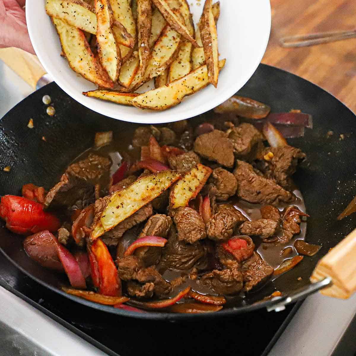 A person transferring crispy steak fries from a white bowl into a large wok filled with simmering Classic Lomo Saltado.