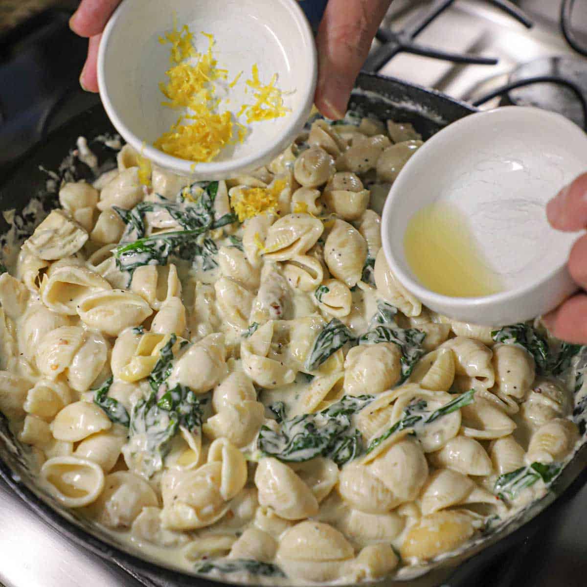 A person transferring lemon zest from a small bowl in one hand and lemon juice in a small bowl in the other hand into a skillet filled with simmering Spinach and Artichoke Dip Pasta.