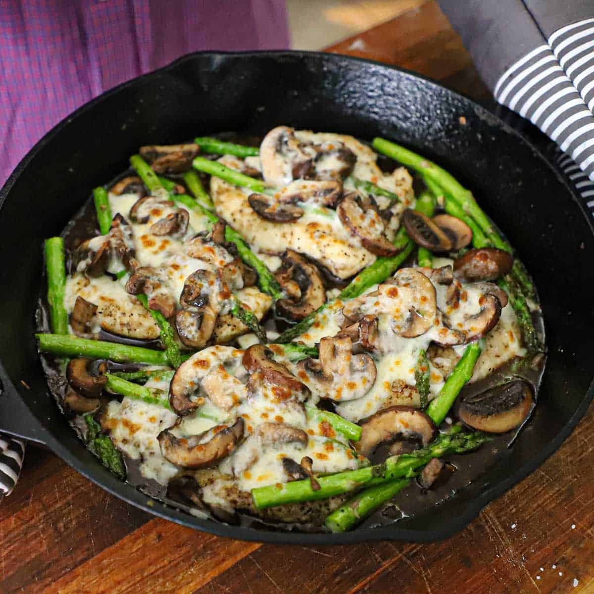 A person wearing oven mitts holding a cast-iron skillet that is filled with a simmering chicken Madeira with mushrooms and asparagus.