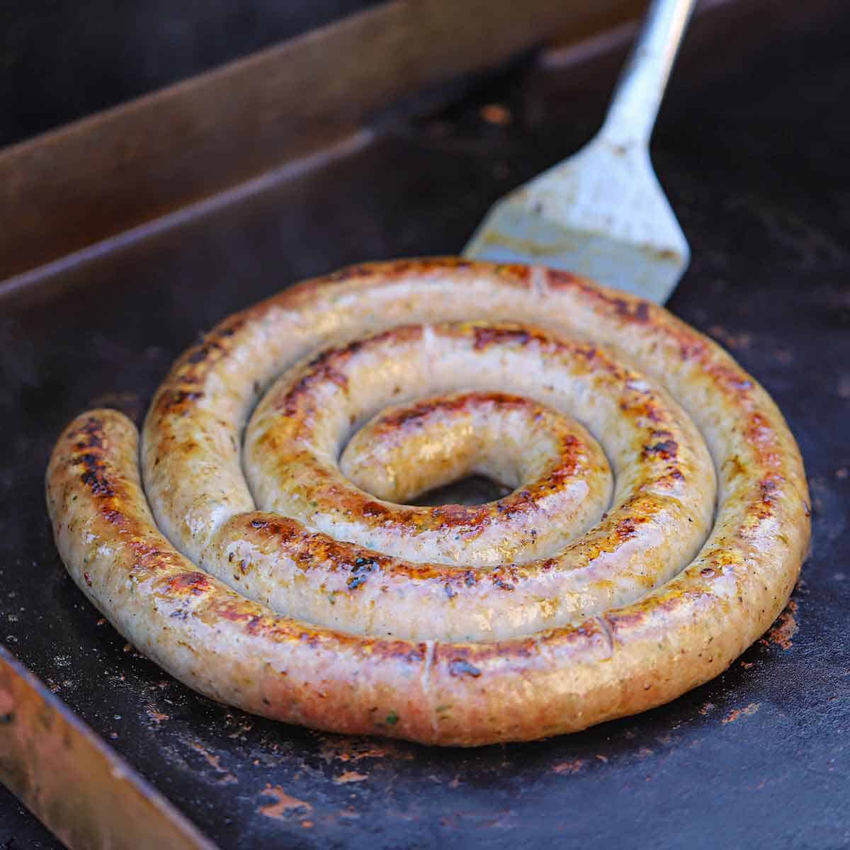 A person using a large spatula to lift up a rope of Italian sausage that is being cooked on an outdoor griddle.