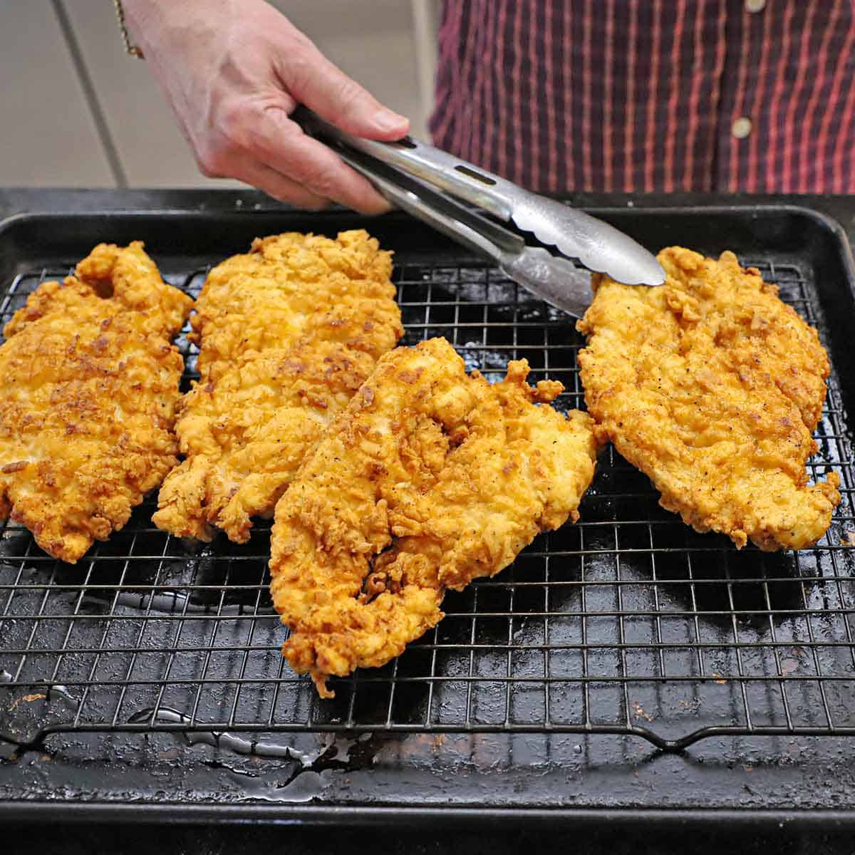 A person using a pair of metal tongs to place cutlets of freshly prepared chicken fried chicken on a wire rack in a baking sheet pan.