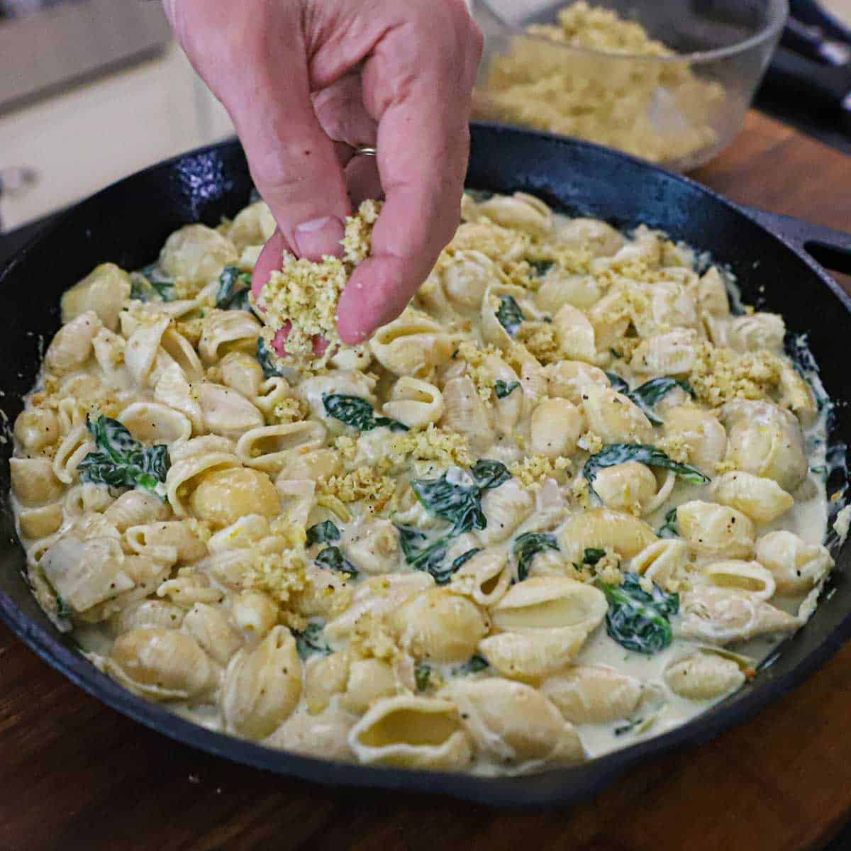 A person sprinkling a Panko breadcrumb and butter mixture over the top of a cast-iron skillet that is filled with Spinach and Artichoke Dip Pasta.