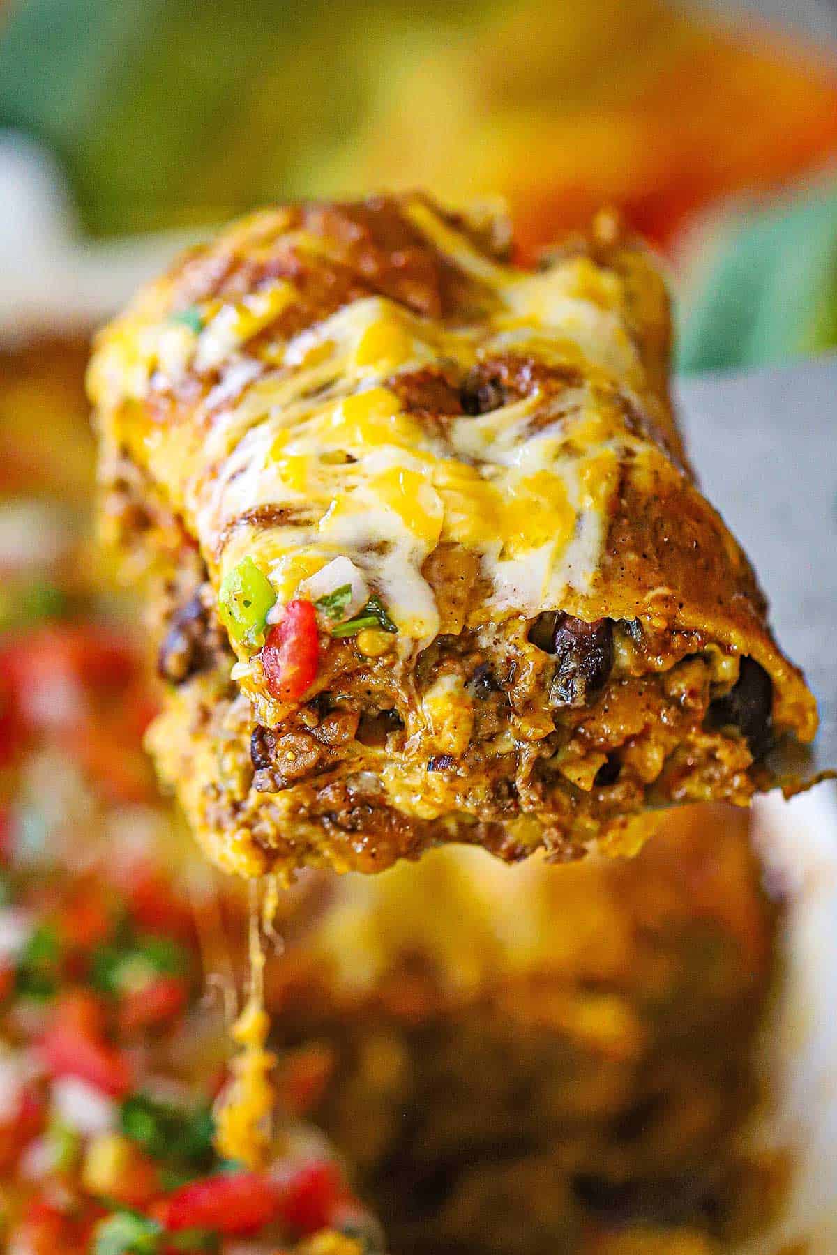 A person using a large metal spatula to raise a large slice of a beef enchilada casserole from a dish filled with the same.
