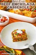 A square slice of beef enchilada casserole on a white dinner plate next to a baking dish filled with the rest of the casserole.