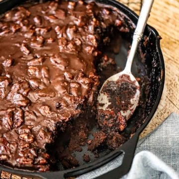 A close-up view of a Fudgy Texas Skillet Cake in a large cast-iron skillet with a portion of it missing and a fudgy serving spoon resting in the pan.