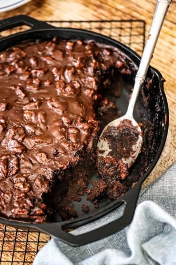 An overhead view of a Fudgy Texas Skillet Cake in a large cast-iron skillet with a portion of it missing and a fudgy serving spoon resting in the pan.