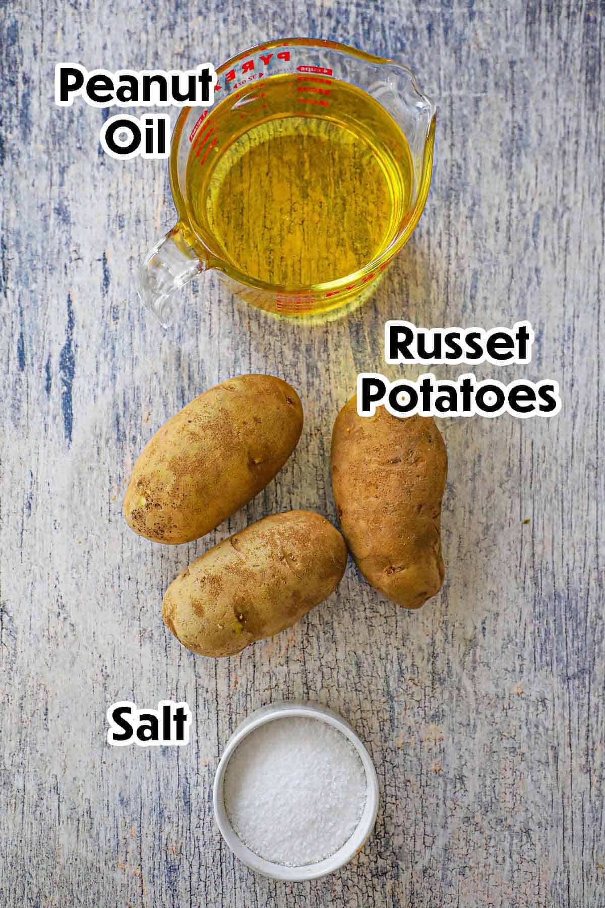 An arrangement of ingredients for homemade French fries on a grey wooden background including russet potatoes, peanut oil, and salt.