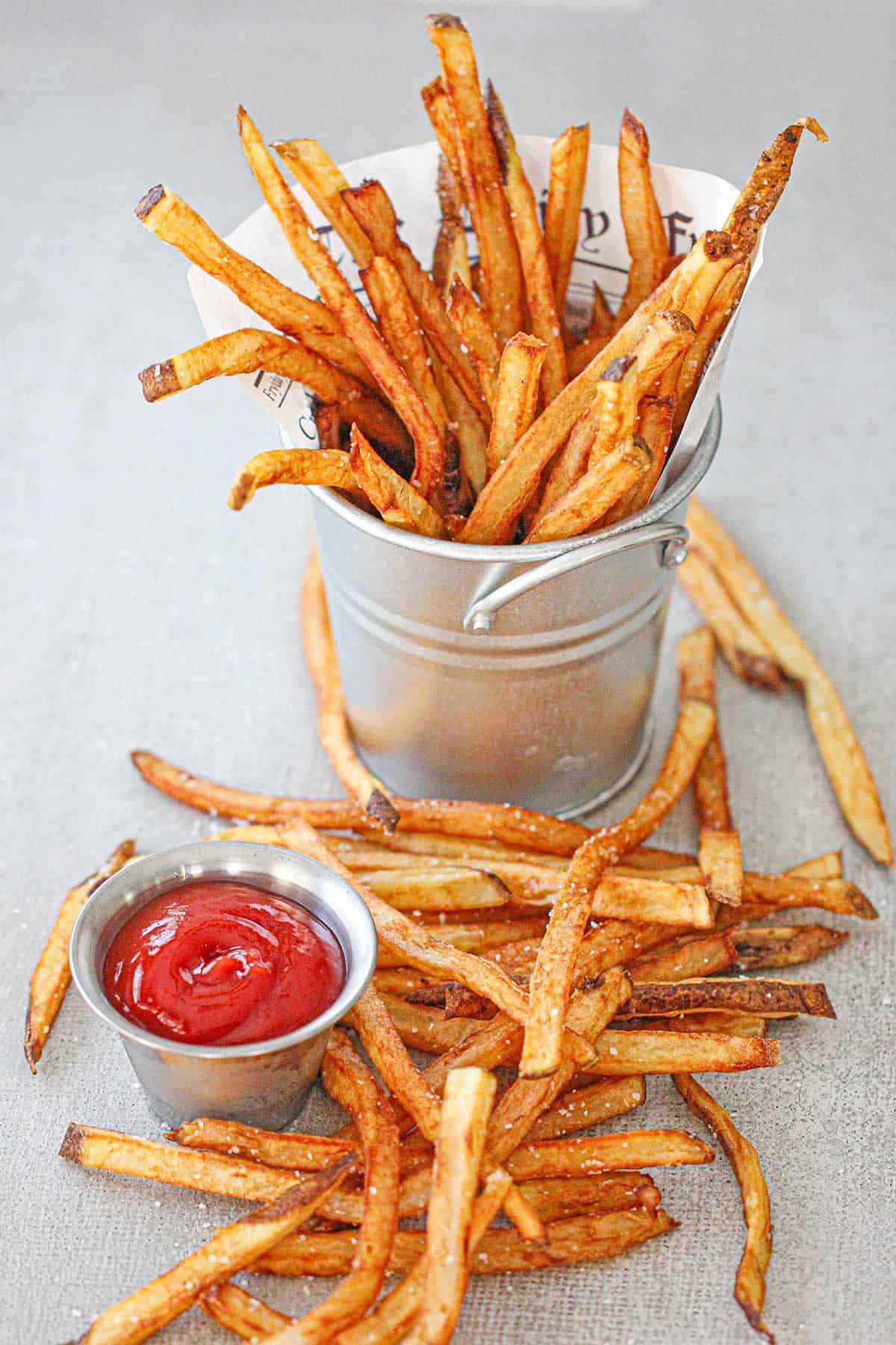 An overhead view of homemade French fried standing upright in a small metal pale next to a pile of fries with a small container of ketchup nearby.