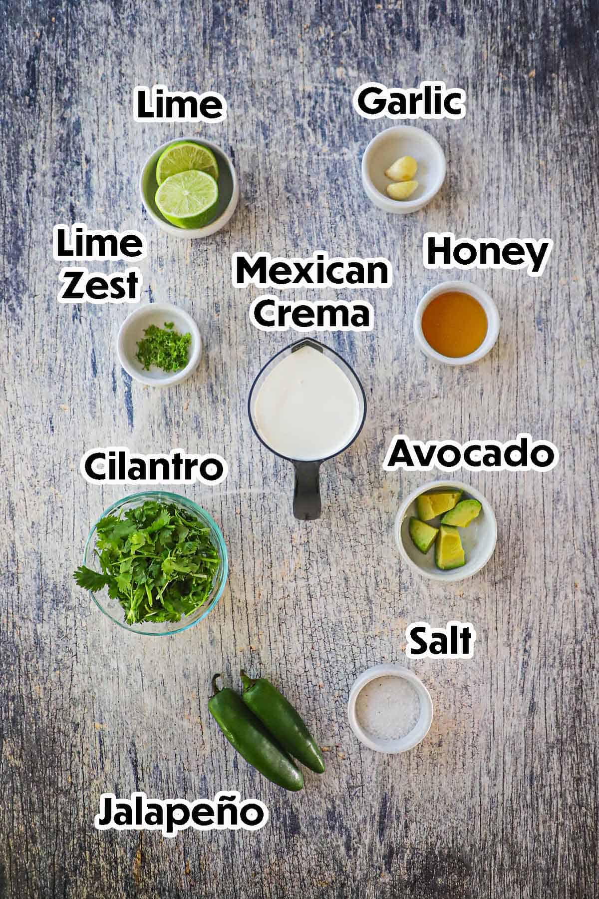 An arrangement of ingredients for Jalapeño-Lime Sauce on a grey wooden background including Mexican crema, cilantro, jalapeños, avocado, limes, lime zest, garlic, and honey.