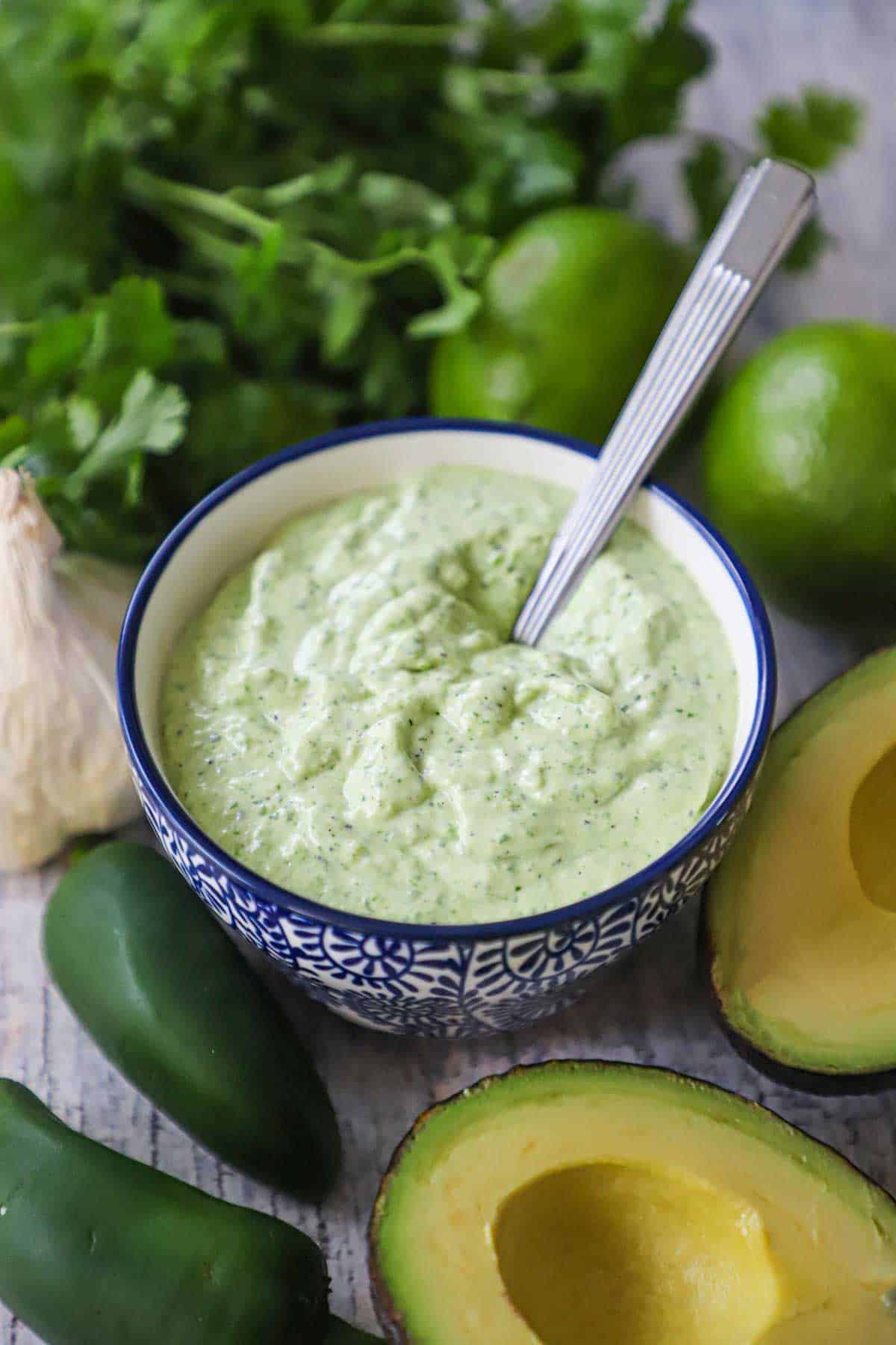 A festive blue and white condiment bowl filled with Jalapeño Lime Sauce with a spoon inserted into it and fresh jalapeños, avocados, and cilantro nearby.