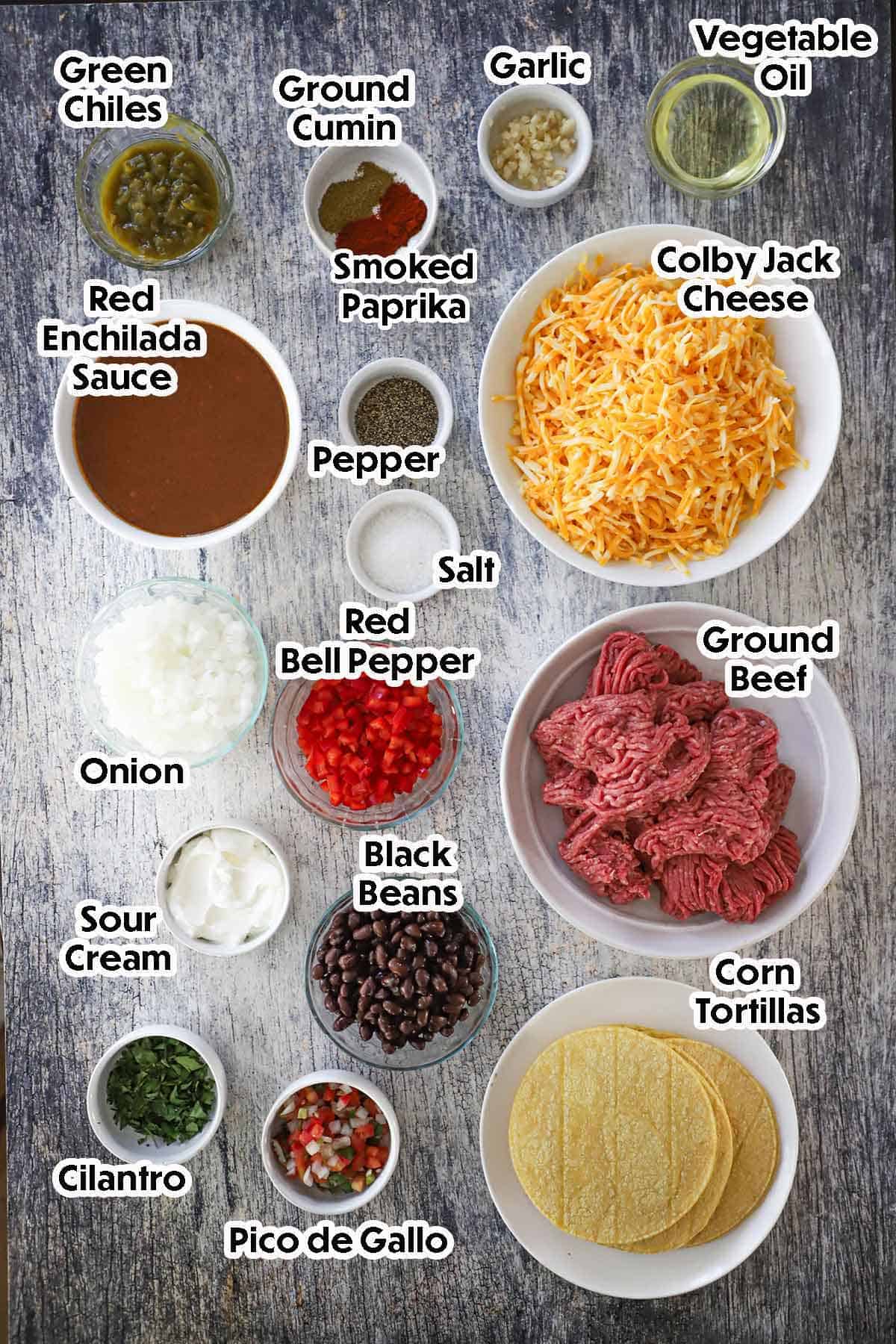 An arrangement of ingredients for beef enchilada casserole on a grey wooden background including ground beef, onions, chopped bell peppers, garlic, enchilada sauce, black beans, corn tortillas, and seasonings.