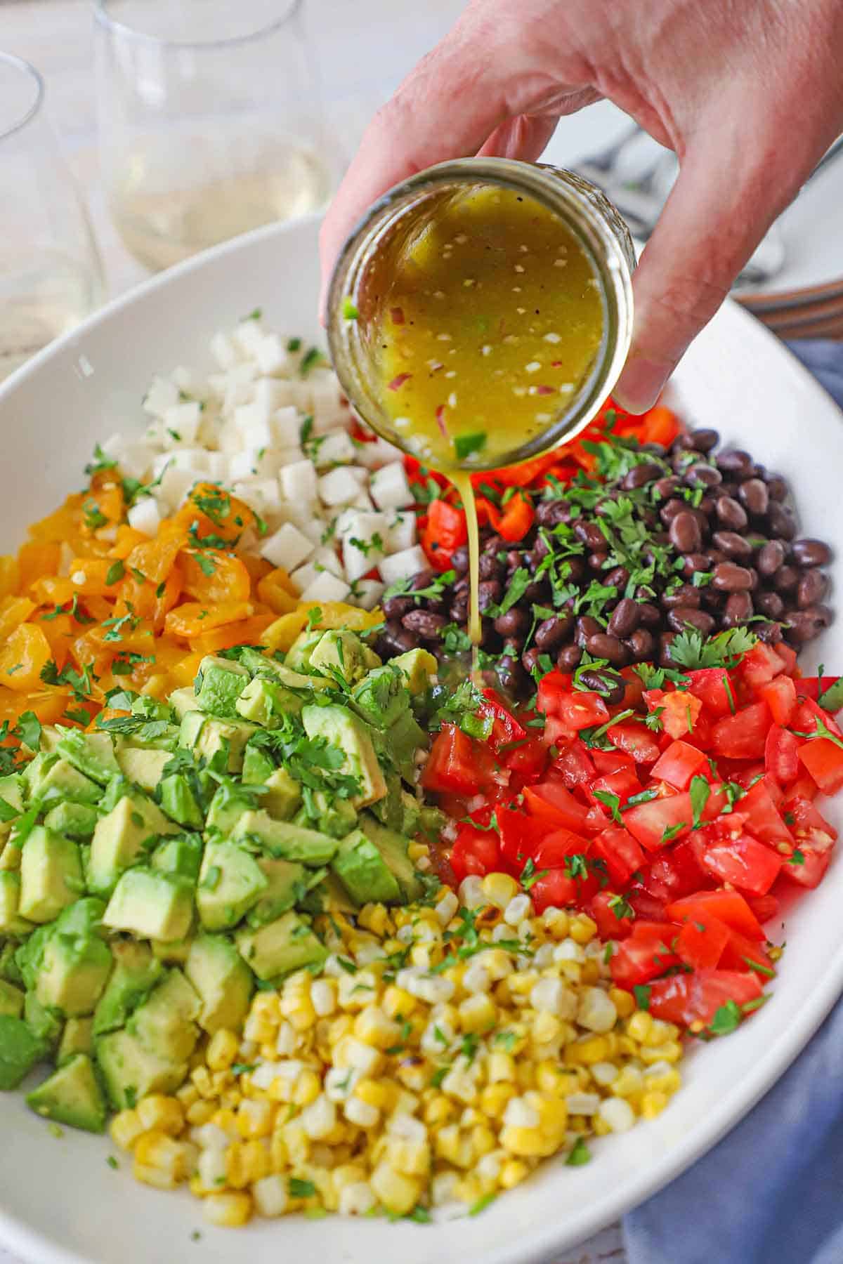 A person pouring a citrus-honey vinaigrette from a small jar over the tops of a deconstructed Southwestern Summer Salad in a large white platter.