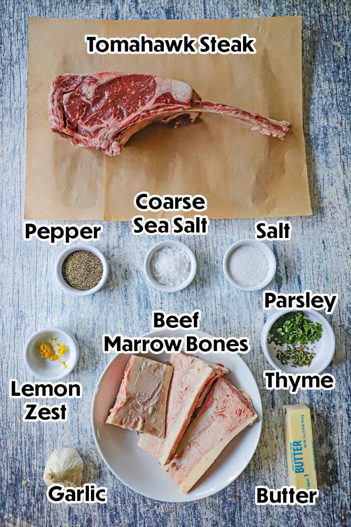 An arrangement of ingredient for tomahawk steak with bone marrow butter on a grey wooden background including a tomahawk steak, beef marrow bones, butter, garlic, lemon zest, and seasonings.