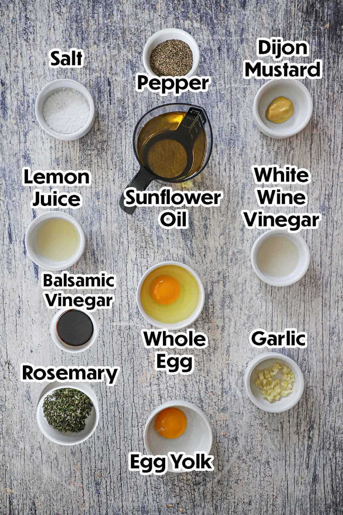 An arrangement of ingredients for Rosemary Garlic Aioli on a grey wooden background including a whole egg, an egg yolk, vinegar, Dijon mustard, lemon juice, oil, salt, chopped rosemary, balsamic vinegar, and minced garlic.