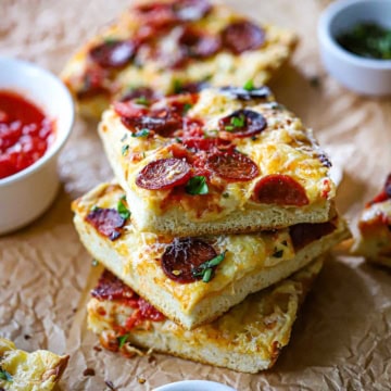 A stack of three square pieces of homemade sheet pan pepperoni pizza on a crumpled brown paper with a small bowl of pizza sauce nearby.
