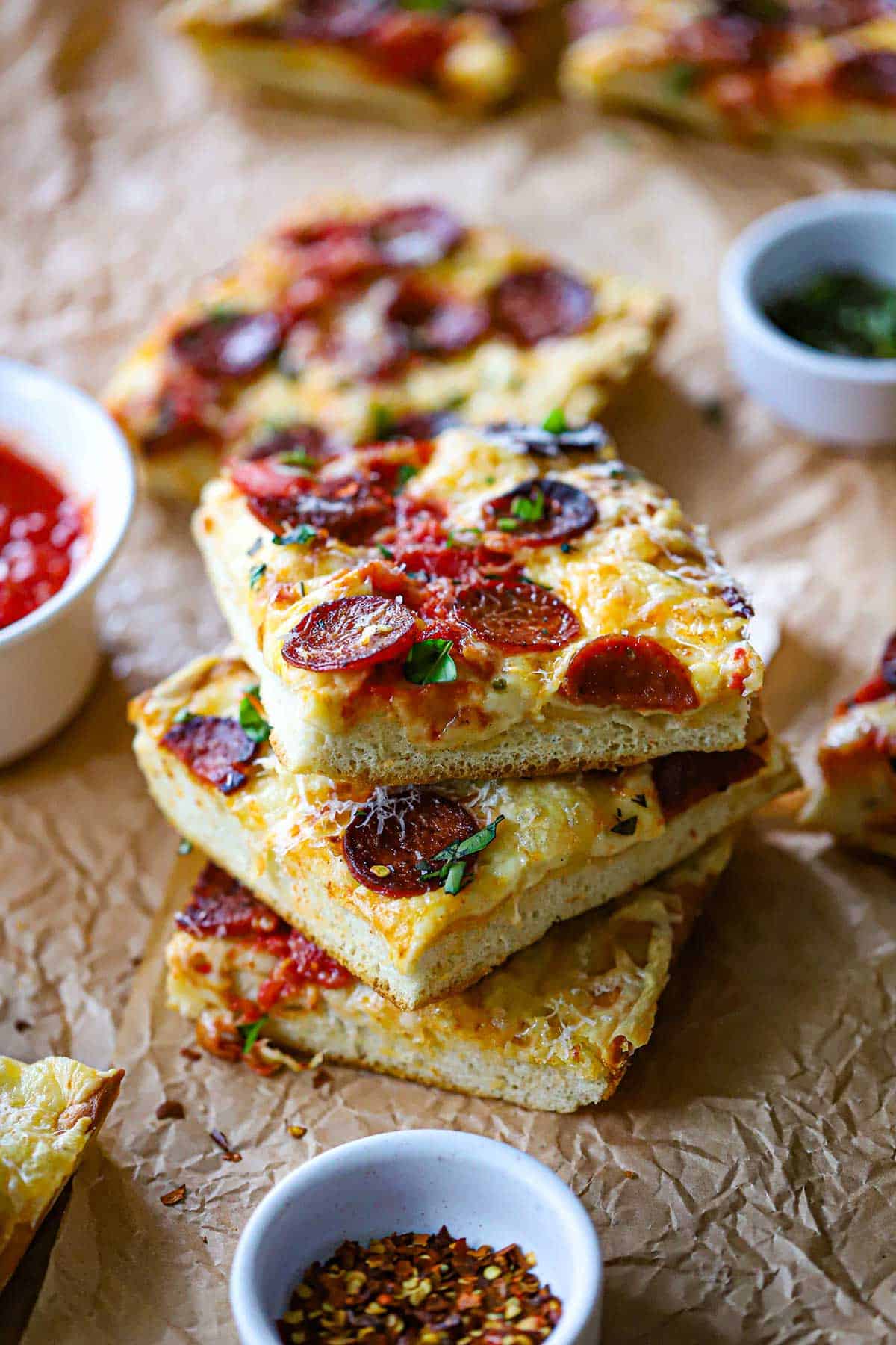 A stack of three square pieces of homemade sheet pan pepperoni pizza on a crumpled brown paper with a small bowl of pizza sauce nearby.