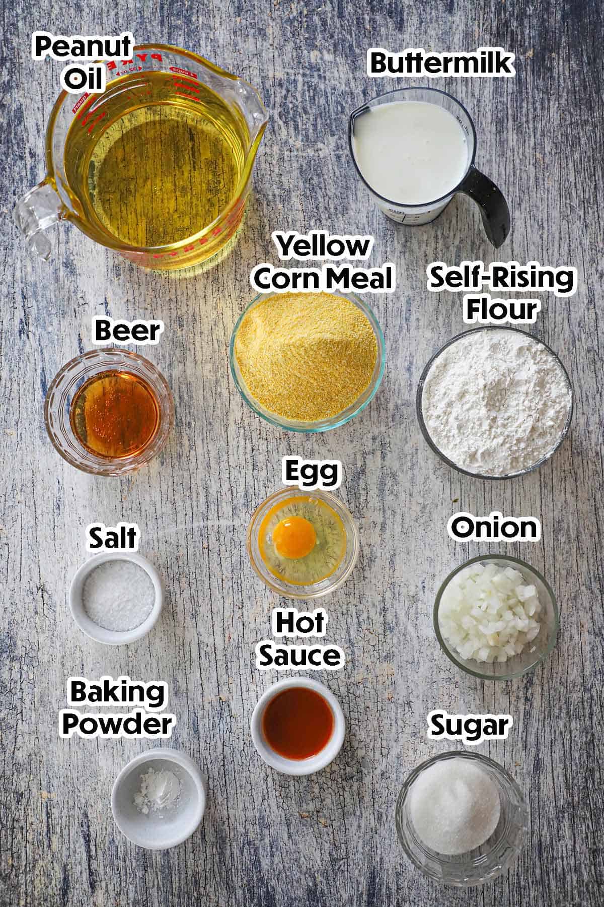 An arrangement of ingredients for Traditional Southern Hush Puppies on a grey wooden background including cornmeal, self-rising flour, buttermilk, beer, egg, onion, sugar, hot sauce, baking powder, and salt.