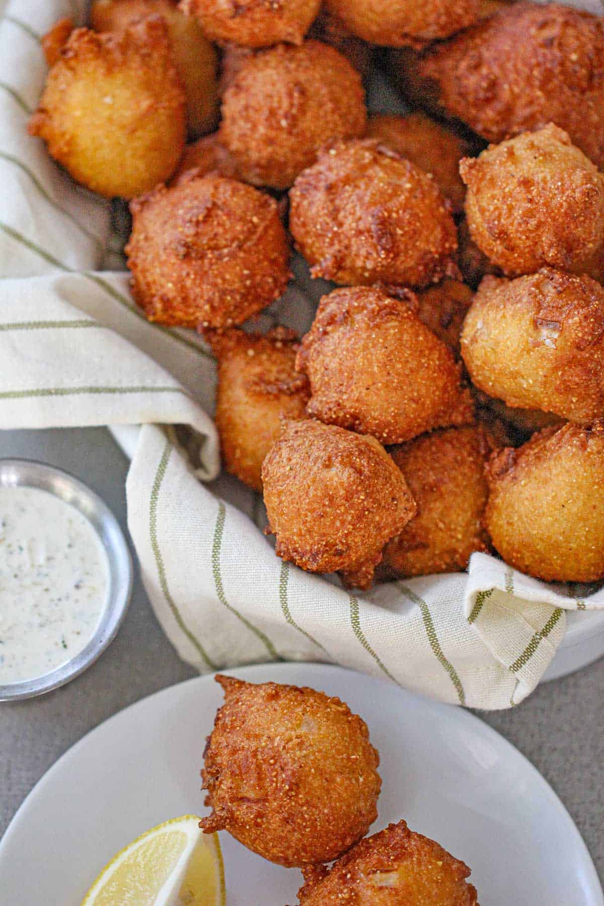 An overhead view of Traditional Southern Hush Puppies pile high in a basket lined with a cloth linen and a plate next to the basket with two more hush puppies and a small bowl of tartar sauce nearby.
