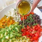 A close-up view of a person pouring a citrus-honey vinaigrette from a small jar over the top of a Southwestern summer salad consisting of piles of corn, tomatoes, avocado, roasted peppers, jicama, and black beans.
