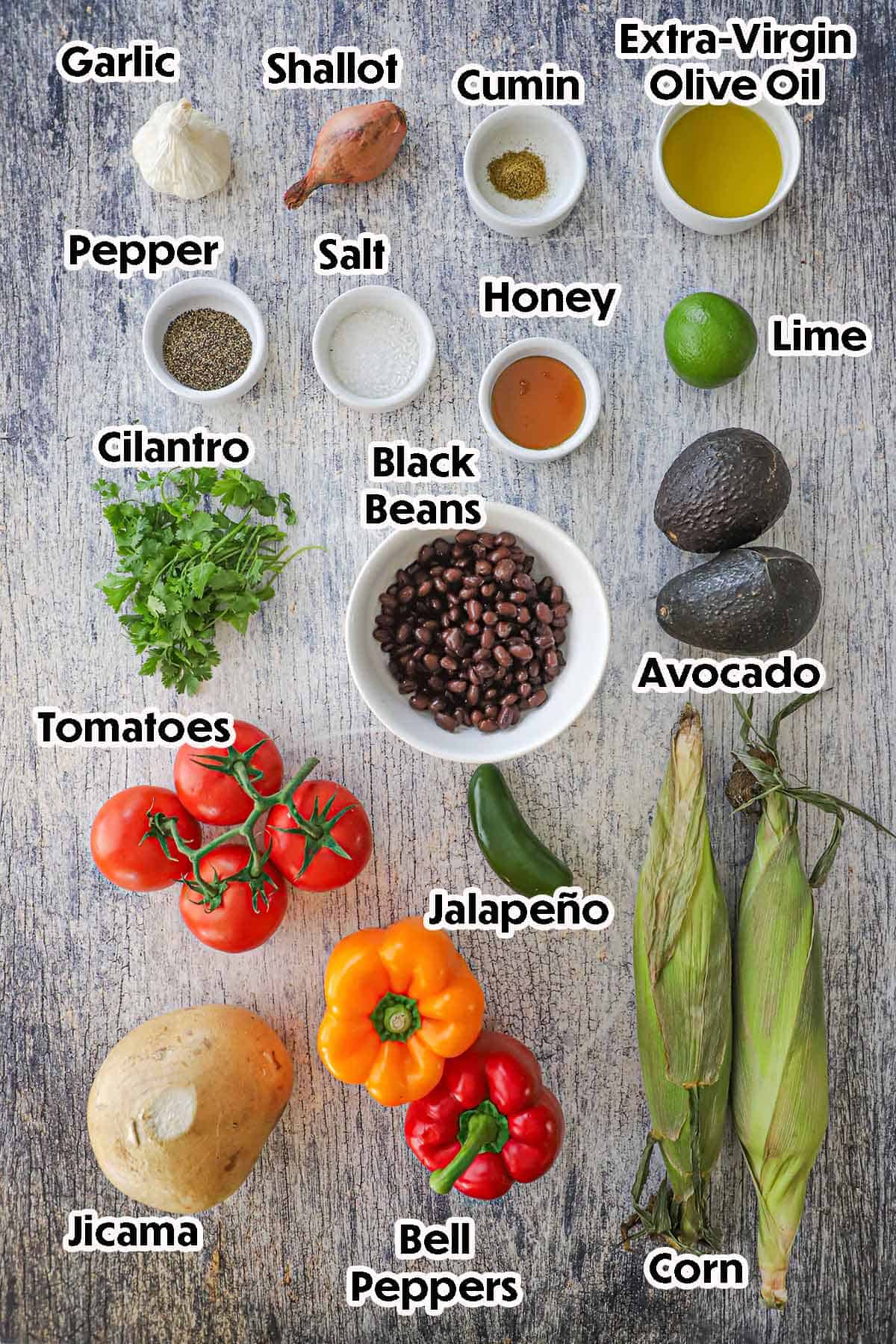 An arrangement of ingredients for a Southwestern Summer Salad on a grey wooden background including ears of corn, bell peppers, jicama, tomatoes, avocado, black beans, cilantro, lime, honey, shallot, garlic, and seasonings.
