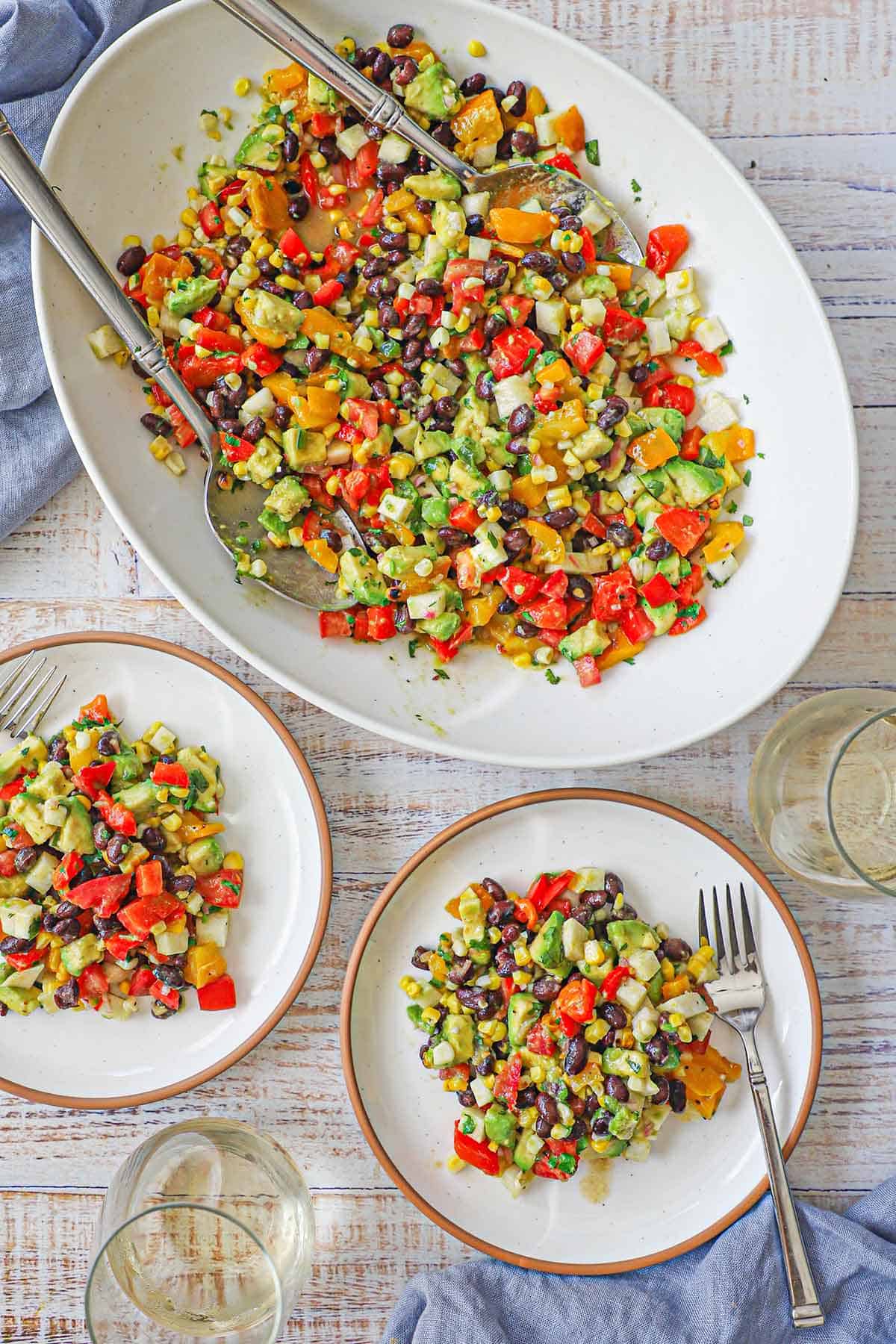 An overhead view of a large white platter filled with a tossed Southwestern Summer Salad next to small salad plates filled with the same.