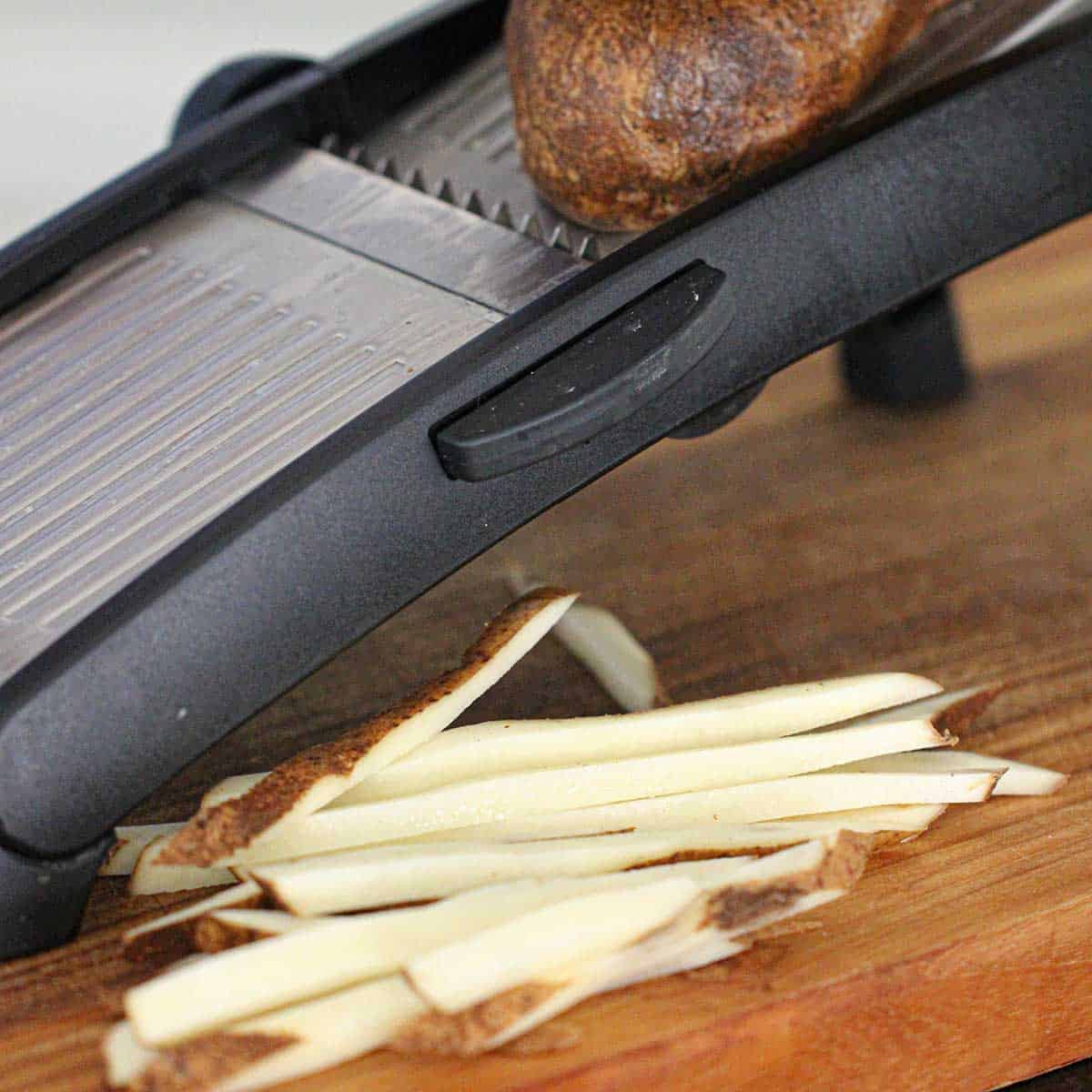 A person using a mandolin to slice a russet potato into strips for homemade French fries.