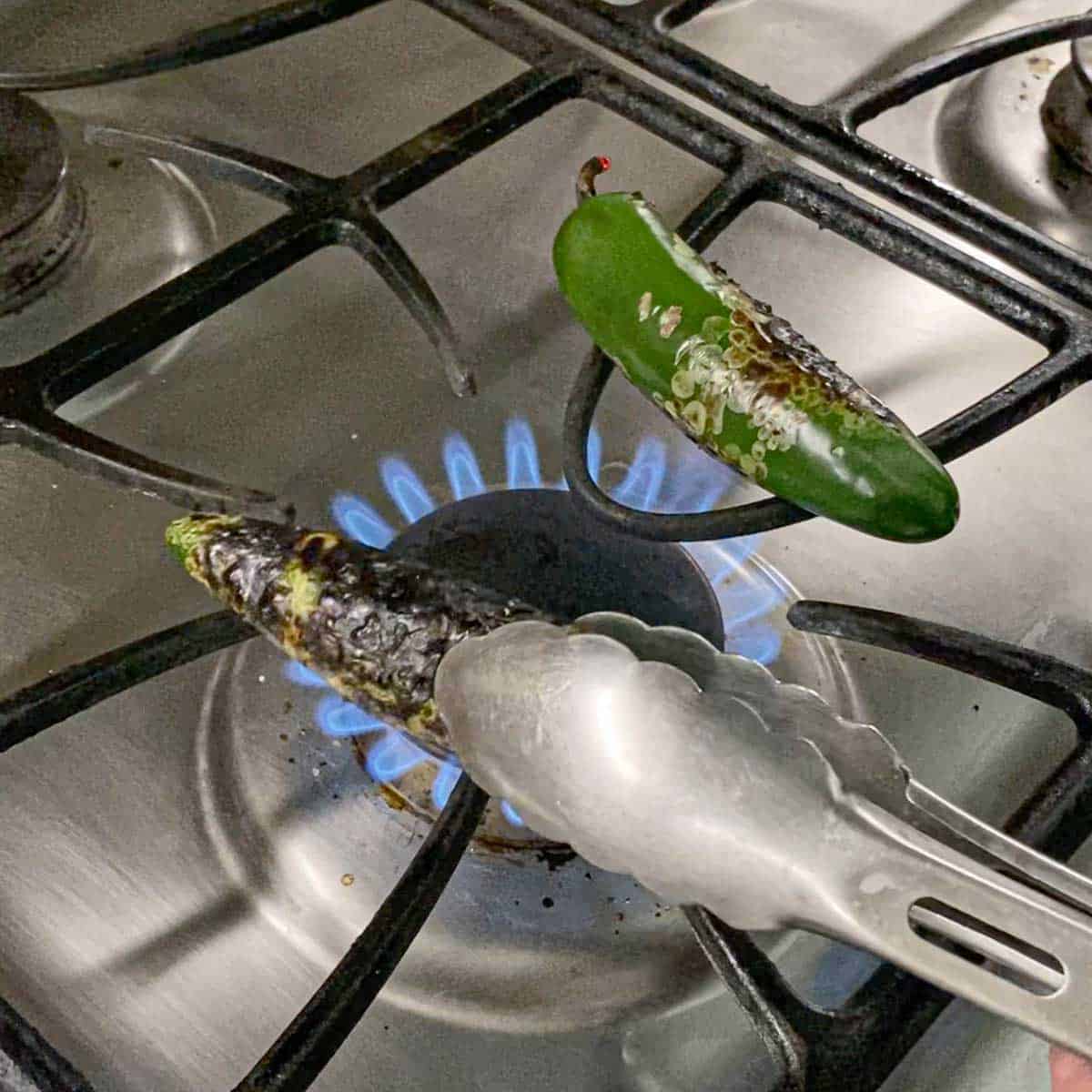 A person using a pair of tongs to char fresh jalapeños over a gas flame on a gas stove.