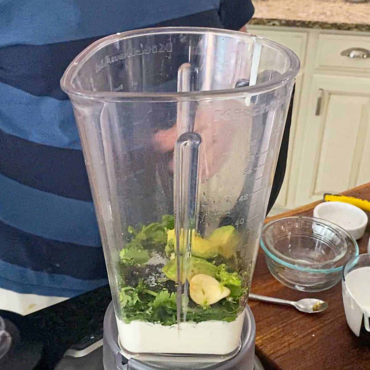 A person standing behind a blender that contains Mexican crema, cilantro, galri, avocado, and seasonings.