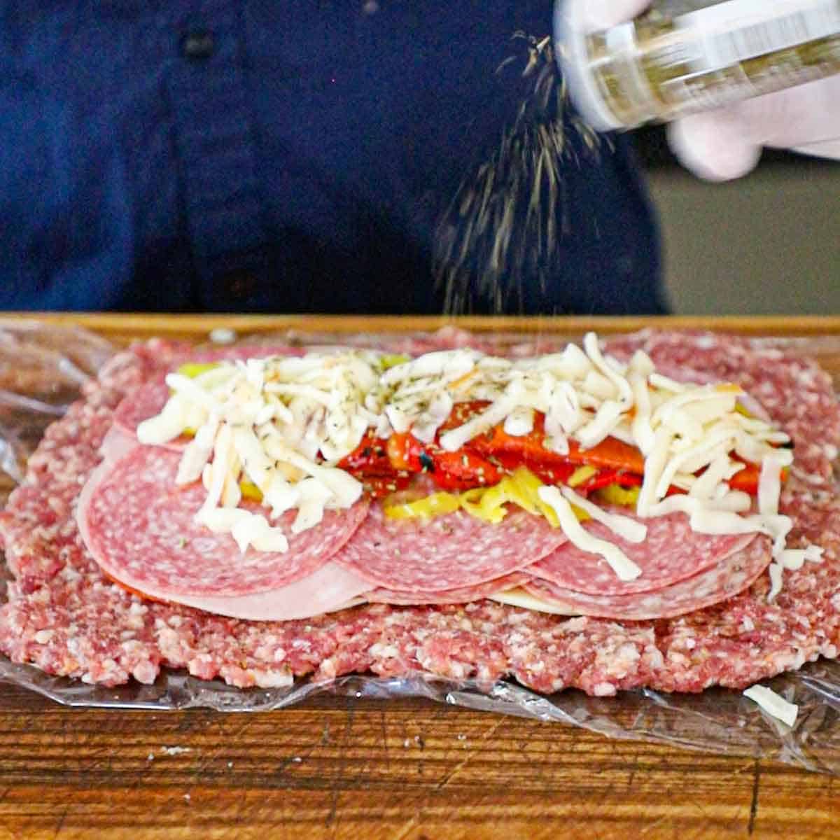 A person sprinkling dried oregano over the top of layers of Italian cold cuts, roasted vegetables, and shredded mozzarella cheese all on a thin layer of Italian sausage resting on a piece of plastic wrap.