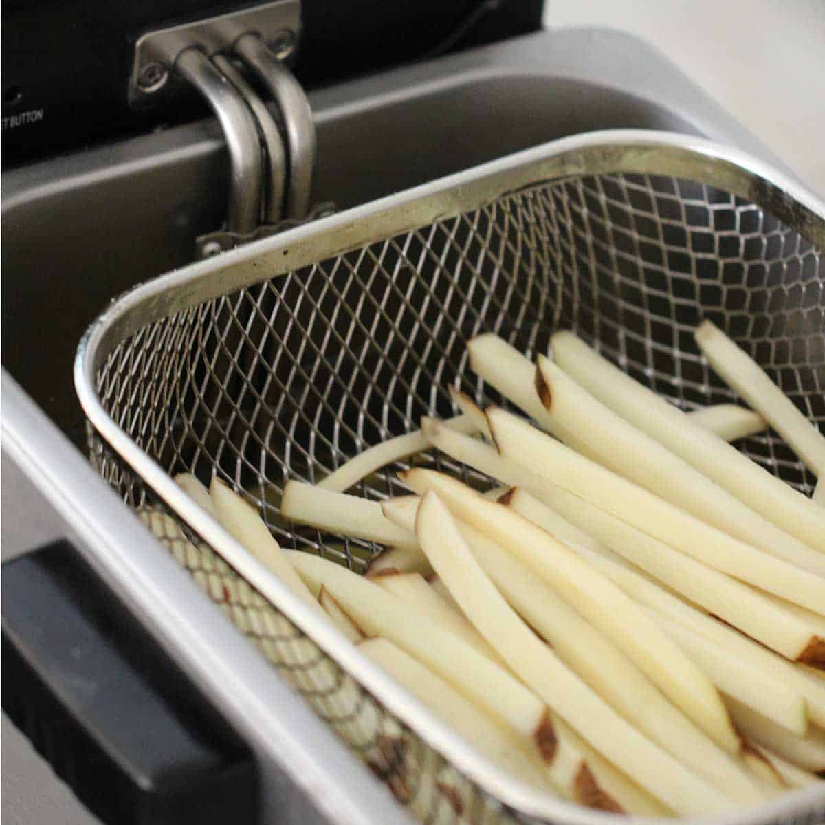A close-up view of a deep-fryer basket filled with uncooked French friers over a vat of hot oil.