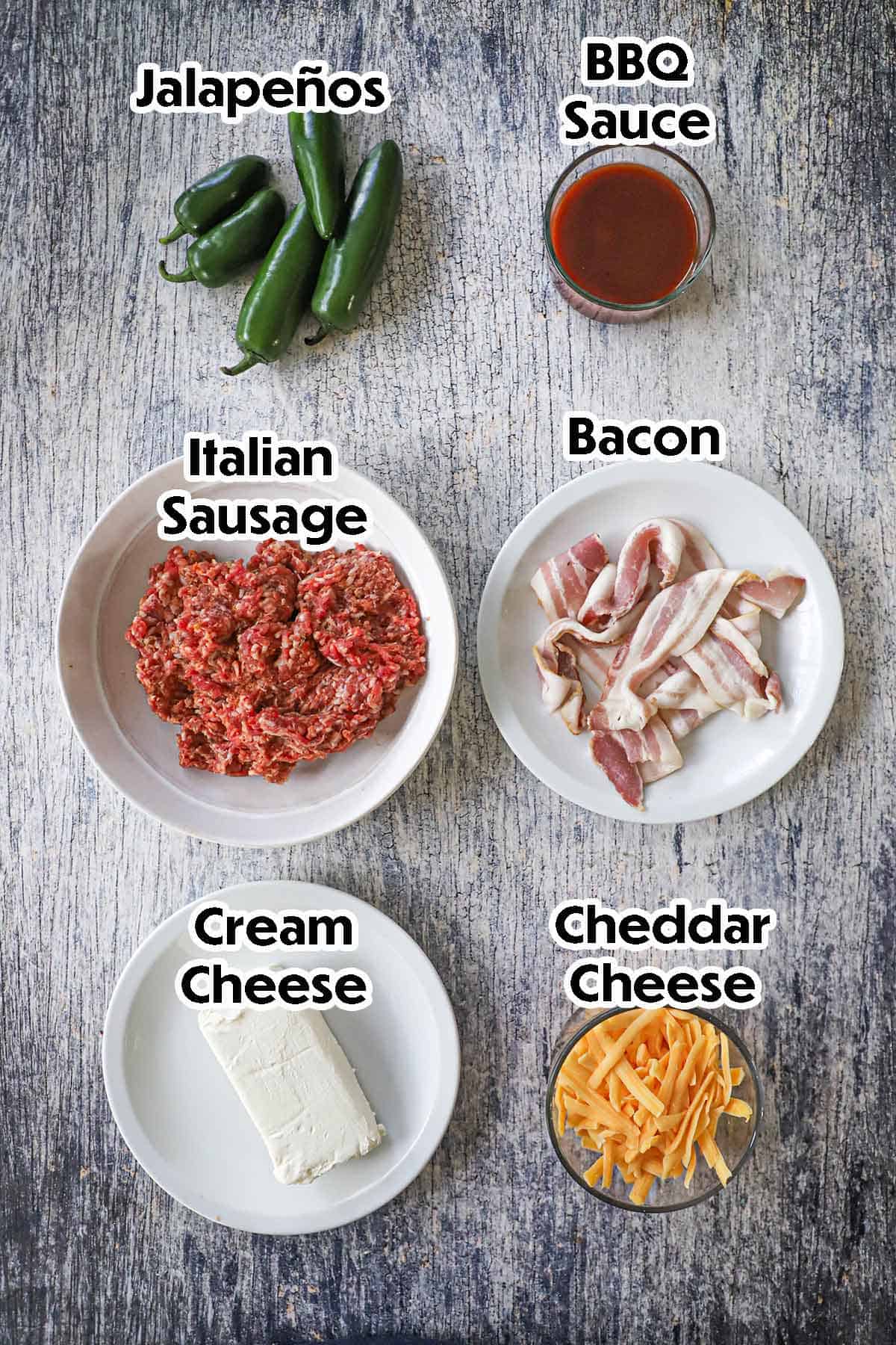 An arrangement of ingredients for smoked armadillo eggs on a grey wooden background including fresh jalapeños, Italian sausage, cream cheese, shredded cheddar, bacon, and barbecue sauce.