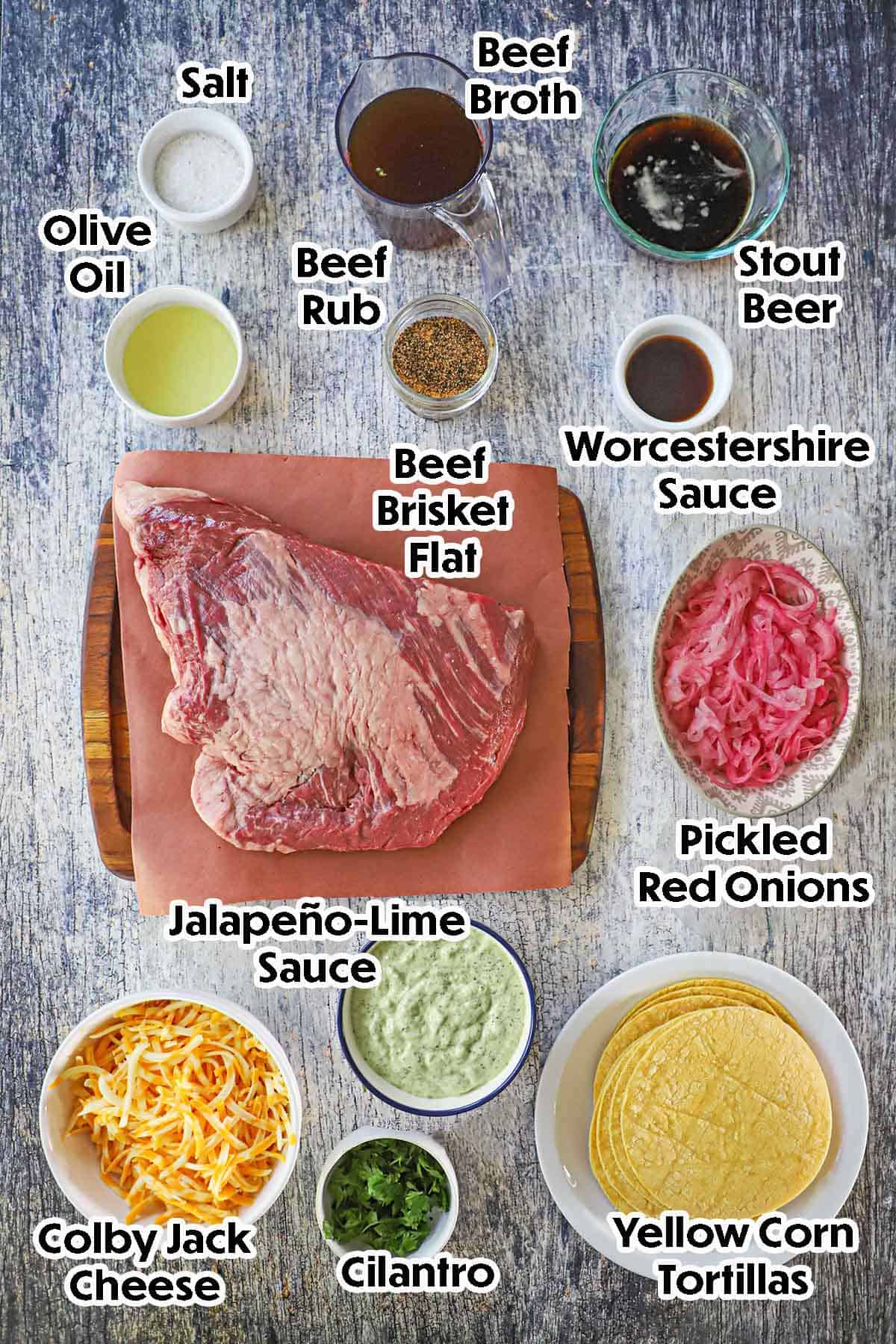 An arrangement of ingredients for baked brisket tacos on a grey wooden background including a beef brisket flat, beef broth, stout beer, Worcestershire sauce, oil, pickled onions, jalapeño-lime sauce, shredded cheese, cilantro, and seasonings.