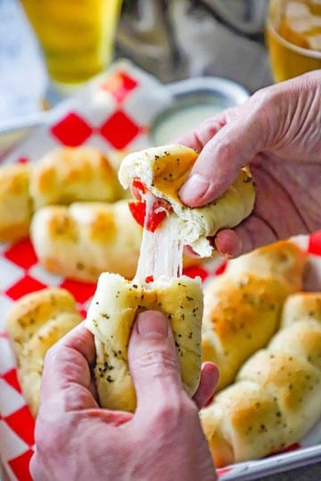 A person using his two hands to pull apart a melt-in-your-mouth pepperoni roll over a trey filled with more pepperoni rolls.