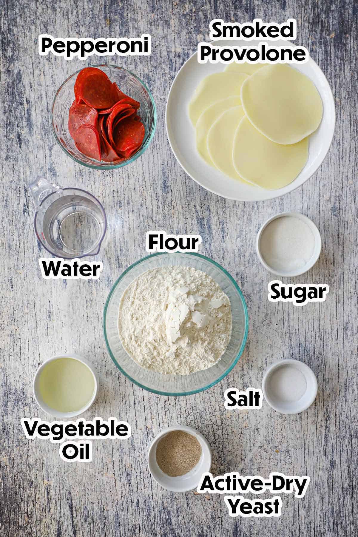 An arrangement of ingredients for Melt-In-Your-Mouth Pepperoni Rolls on a grey wooden background including flour, water, sugar, salt, oil, yeast, pepperoni slices, and sliced smoked Provolone.