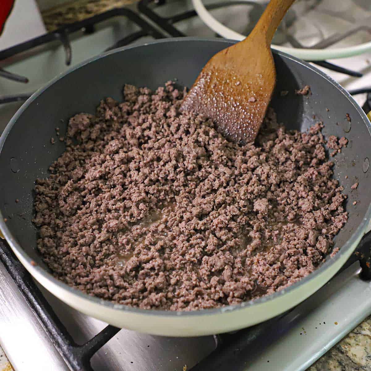 A large non-stick skillet filled with fully cooked ground beef with a large wooden spatula off to the side of the skillet.